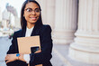 © BullRun - Half length portrait of cheerful dark skinned businesswoman in formal wear holding smartphone and folder smiling at camera.Successful african american student of faculty of law with crossed hands