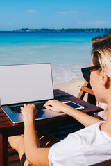 Wall Mural - Back view of young woman enjoying distance work on beautiful ocean beach typing on laptop computer with mock up screen,female blogger watching video on netbook and chatting with followers on weekends