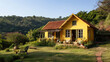 © Karolis - A bright yellow house is set in a lush, hilly landscape. The garden is well-maintained with outdoor seating, and the sky is clear, suggesting a peaceful day.