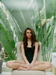 © SHOTPRIME STUDIO - Young woman sitting cross-legged on a chair, wearing a black swimsuit, looking pensive, with large green leaves in the background.