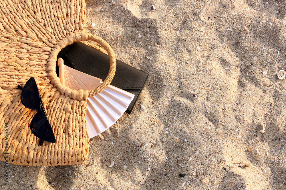 Stylish sunglasses and straw bag with wallet on sandy background