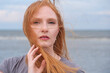 © ullision - Woman with red hair stands by the sea, hair blowing across her face. gazes directly at camera with a serious expression, touching her hair with one hand. sky and ocean form the background.