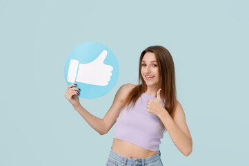 Pretty young woman holding like icon and showing thumb-up on blue background