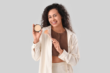 Wall Mural - Beautiful young happy African-American woman with makeup brush and powder on grey background