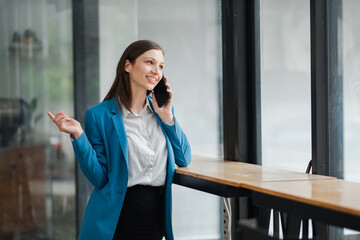 Wall Mural - Professional businesswoman in a blue blazer smiling while talking on a smartphone in a modern office with large windows.