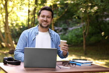 Canvas Print - Smiling freelancer drinking coffee at table with laptop outdoors. Remote job