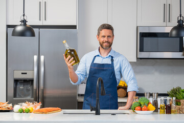 Wall Mural - Handsome man in cook apron cooking with bottle olive oil in kitchen. Portrait of middle aged man in chef apron hold bottle olive oil in kitchen.