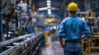 © atipong - Factory worker in blue shirt and yellow hard hat inspects machine in busy factory setting, deep in thought.