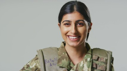 Wall Mural - Smiling young female soldier in uniform wearing body armour in front of plain studio background - shot in slow motion