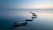 © lucegrafiar - A serene image of large stepping stones leading across a calm sea during sunrise.