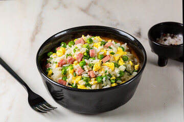 Wall Mural - Cantonese Fried Rice and egg omelette with green pea, ham steak, spring onion, ribe long rice in a beige bowl on a white marble table, served with fork. Eurasian cuisine.