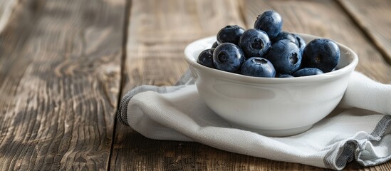 Poster - A small white bowl filled with organic blueberries sits on a wooden table next to a kitchen towel, with copy space in the image. Summer berries; fresh forest fruit.