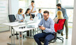 © Bojan - Handsome businessman sitting at his workplace, taking a break while using his mobile phone, with his colleagues working in the background.