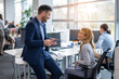 © Bojan - Businessman sitting on the desk while casually chatting with his female colleague who is seated at her workstation in the office.