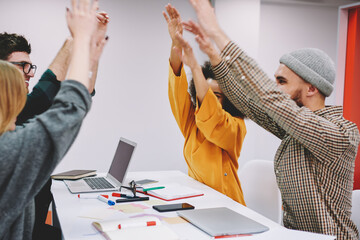 Fototapete - Cheerful deversity group of male and female designers with their hands raised celebrating accomplished project sitting at desktop.Successful multicultural students have won during brainstorming