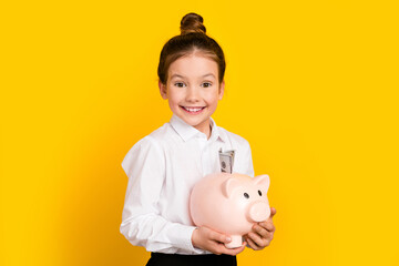 Portrait of cute schoolgirl hold money bank pig wear uniform isolated on yellow color background
