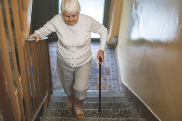 Poster - Elderly woman walking up the stairs leaning on a wooden cane
