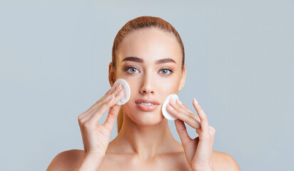 Wall Mural - This image shows a young woman with a neutral expression applying cotton rounds to her face. She is standing in front of a plain light blue background in a studio setting.