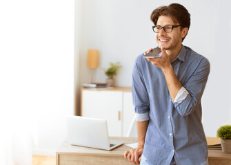 Poster - A man wearing glasses stands in his office, smiling while using a voice assistant on his phone. He is leaning against a desk with a laptop and other office supplies.