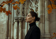 © krit - A sophisticated woman in a black coat stands against an ornate stone building with autumn leaves in the foreground.