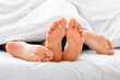 © Prostock-studio - This image shows the feet of two people resting under white sheets in a bed. The feet are positioned close together, suggesting intimacy and comfort