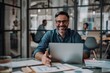 © ammad - A smiling man in glasses sitting at desk with laptop holding pen looking into camera Professional stress free Workspace Concept