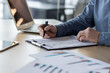 © Tetiana - Close-up photo of the hands of a young businessman in a blue shirt sitting at a desk, holding a pen, working with data and documents, making notes and signing