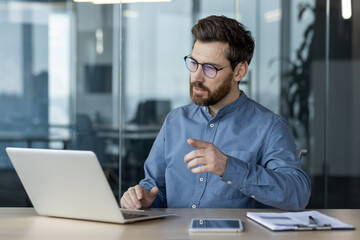Wall Mural - Serious and focused young businessman in glasses and blue shirt sitting in the office at the table and having a video call, conference and online meeting