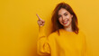 © vitor - Mulher jovem e atraente sorrindo e apontando em pé contra uma parede amarela, fundo isolado, fêmea adulta em suéter amarelo casual, sobre um ambiente moderno colorido