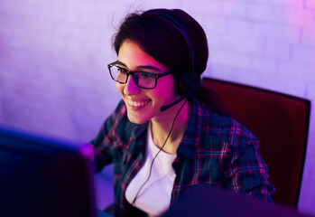 Poster - A young woman with dark hair is wearing glasses and a headset while sitting in front of a computer. She is smiling and looking at the screen. The room is lit in a purple and pink glow