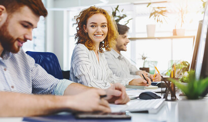 Poster - Cheerful redhead girl smiling at camera, working at coworking with other people, empty space