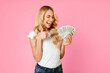 © Prostock-studio - A young woman with long blonde hair is smiling and giving a thumbs up while holding a fan of cash in her other hand. She is wearing a white t-shirt and blue jeans and is standing against pink