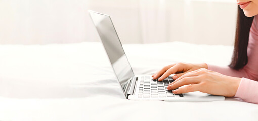 Poster - A woman is sitting on a bed, focused on using a laptop computer. She is typing and browsing online content, cropped, panorama