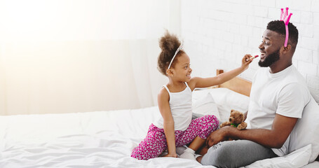 Wall Mural - African American little girl, wearing a white tank top and pink floral pants, is applying lipstick to her fathers lips. They are both sitting on a white bed, and the girl is smiling.