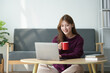 © amnaj - Smiling woman working on a laptop while holding a red coffee mug, seated at a wooden table in a cozy home setting.