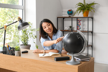 Wall Mural - Asian Geography teacher with globe sitting at table in classroom