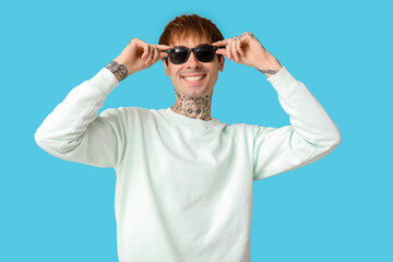 Wall Mural - Happy handsome young tattooed man with sunglasses on blue background