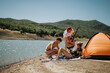 © qunica.com - Group of friends camping by the lake with a guitar, enjoying a sunny day outdoors. They are relaxing by their tent and having fun together.