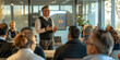 © Pam - Business professional in sweater vest presenting to diverse group in modern glass-walled office. Engaged audience in casual outfits listening to business strategy
