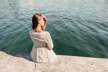 Wall Mural - A woman is sitting on a ledge by a body of water. She is wearing a white dress and she is looking out at the water. The scene is peaceful and serene, with the woman taking a moment to enjoy the view.