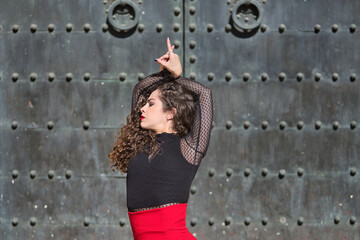 Canvas Print - Portrait of young, beautiful, brunette woman in black shirt and red skirt, dancing flamenco with old, black metal door in the background. Flamenco concept, dance, art, typical Spanish.