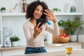 Wall Mural - Beautiful woman doing a videocall with her smartphone while drinking a cup of tea in the kitchen at home.