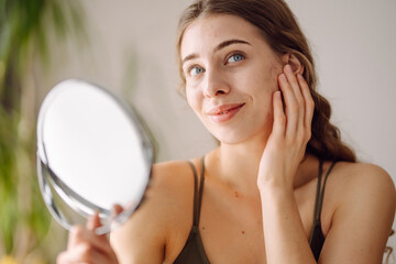 Wall Mural - Young woman with problem skin looking into mirror. Concept of acne therapy, scars, inflammation on face, problem skin. Beauty care.