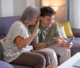 Video call concept. Smiling handsome boy and his grandmother sitting together on sofa using telephone smartphone technology for online webcam connection