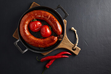 Wall Mural - Fried pork sausage and grilled tomatoes on a black cast iron frying pan on a stone table with copy space