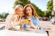 © maxbelchenko - Two young women, sisters, best friends showing news social media photos on smart phone cellphone sitting in cafe and drinking coffee during breakfast. Fashion, beauty, blogging, tourism.