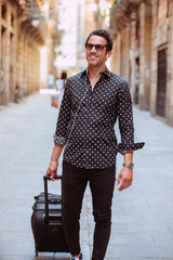 Portrait of a traveling smiling handsome caucasian business man tourist, pulling suitcase, wears stylish clothes and sunglasses, listening music with earphones, walking at the street during travel.