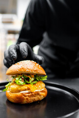 Wall Mural - A close-up of a chef in a black uniform garnishing a gourmet burger with greenery on a black plate, showcasing culinary art and precision