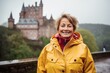 © Markus Schröder - Portrait of a jovial woman in her 60s sporting a waterproof rain jacket over historic castle backdrop