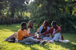 © DimaBerlin - African american students girls and guys having picnic rest on grass in park sharing latest news. Tired group black friends relaxing after university test, taking lunch break sitting on lawn in campus